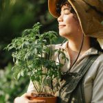 Cultivating Mindfulness - Content ethnic female grower in panama hat with potted plant and closed eyes in summer garden