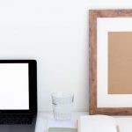 Home Organization - Laptop with blank screen and glass of water placed on white desk near empty frame and blank notepad against white wall