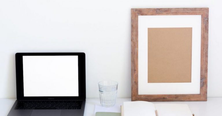 Home Organization - Laptop with blank screen and glass of water placed on white desk near empty frame and blank notepad against white wall