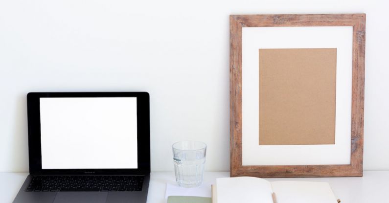 Home Organization - Laptop with blank screen and glass of water placed on white desk near empty frame and blank notepad against white wall
