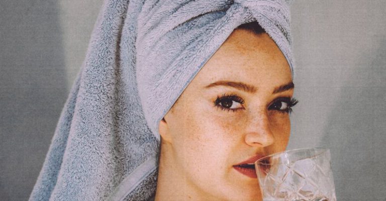 Natural Skincare - Young woman with hair wrapped with towel and bared shoulders drinking water and looking at camera